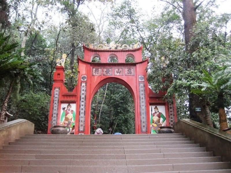 Tour Du Lịch Đền Hùng Cổng vòm màu đỏ được trang trí công phu với các ký tự châu Á ở phía trên, hai bên là những hàng cây cao. Cầu thang dẫn lên trên, với những bức tranh tường đầy màu sắc và phông nền là khu rừng tươi tốt.