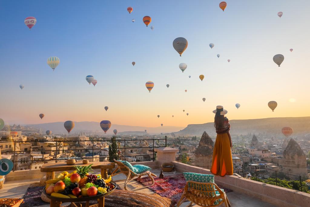 Tour Du Lịch Thổ Nhĩ Kỳ 8 Ngày 7 Đêm Một người phụ nữ trong bộ váy dài thướt tha và đội mũ đang chiêm ngưỡng vô số khinh khí cầu bay lơ lửng trên bầu trời Cappadocia lúc bình minh. Phía trước là một sân hiên đầy màu sắc với trái cây và ghế ngồi.