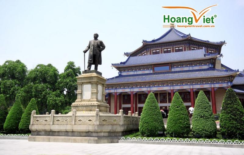 Tượng đồng trước hội trường kiến trúc cổ mái ngói xanh, cột đỏ và hàng cây cắt tỉa.