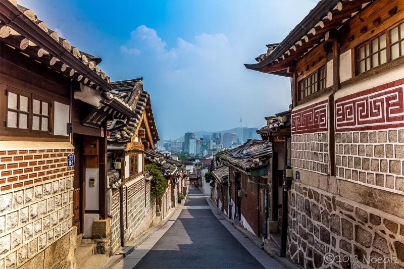 Con hẻm nhỏ giữa những ngôi nhà hanok tường đá, phía xa là các tòa nhà cao tầng của Seoul.