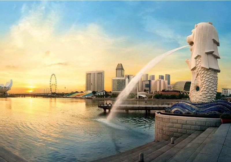 Cảnh hoàng hôn trên vịnh Marina với bức tượng Merlion phun nước nổi tiếng ở phía trước. Đường chân trời nổi bật với những tòa nhà hiện đại và vòng đu quay.