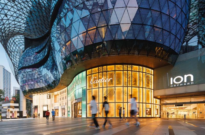 Mặt tiền kính hiện đại của trung tâm mua sắm ION Orchard lúc hoàng hôn. Ánh sáng ấm áp từ các cửa hàng sang trọng, bao gồm cả Cartier, và hình ảnh những người mua sắm đi ngang qua mờ ảo.