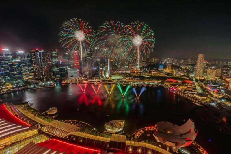 Màn trình diễn pháo hoa rực rỡ trên bầu trời đêm của thành phố, phản chiếu sắc màu xuống mặt nước. Ánh đèn thành phố lung linh bao quanh vịnh, tạo nên một bầu không khí lễ hội.