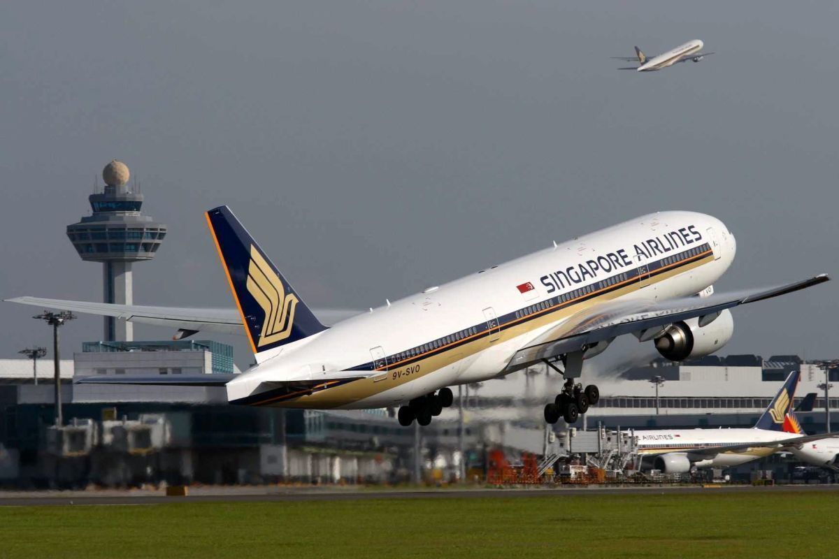 Máy bay của hãng Singapore Airlines cất cánh từ sân bay, phía sau là tháp điều khiển và một máy bay khác đang bay lên phía trên. Bầu trời trong xanh, cho thấy chuyến đi sẽ diễn ra suôn sẻ.
