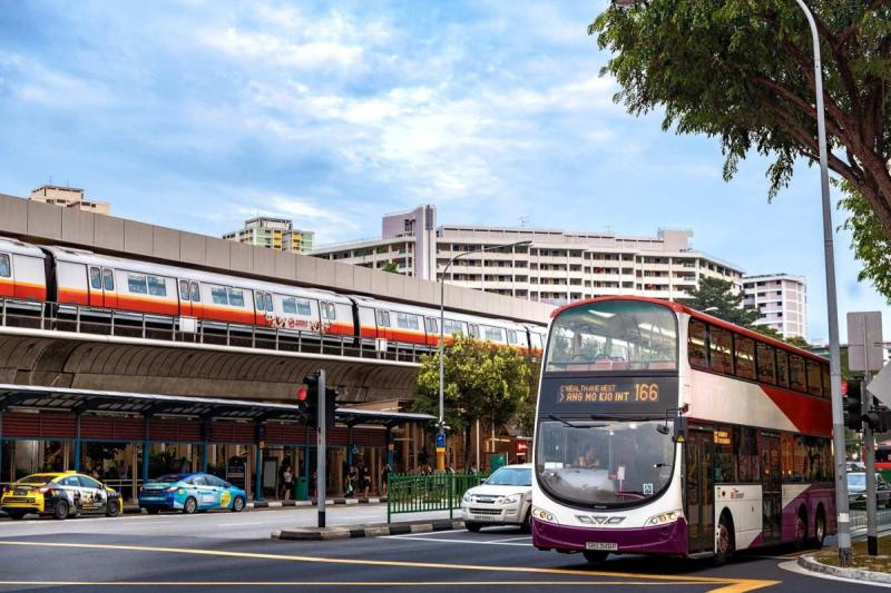 Một con phố đô thị nhộn nhịp với xe buýt hai tầng đang chạy qua, taxi chờ sẵn và đường ray xe lửa trên cao ở phía xa. Bầu trời xanh và cây cối càng làm tăng thêm vẻ sống động.