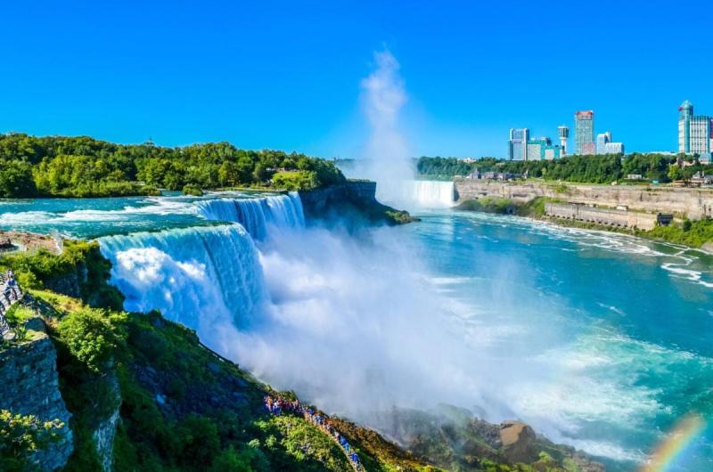 Khung cảnh hùng vĩ của thác Niagara với những dòng thác mạnh mẽ đổ xuống dưới bầu trời xanh trong vắt. Cây cối xanh tươi bao quanh khu vực, trong khi những tòa nhà hiện ra ở phía xa.