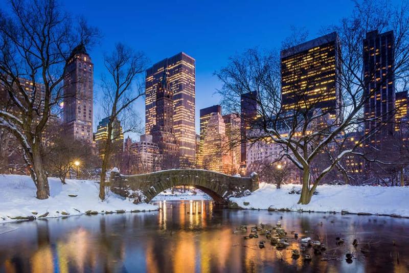 Công viên Central Park phủ đầy tuyết trắng lúc hoàng hôn nổi bật với cây cầu đá bắc qua một hồ nước tĩnh lặng, bao quanh là những hàng cây trơ trụi. Những tòa nhà chọc trời được thắp sáng rực rỡ chiếu rọi bầu trời mùa đông.