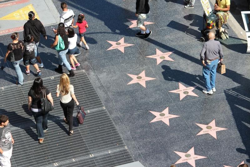 Người đi bộ tản bộ dọc theo Đại lộ Danh vọng Hollywood, với những ngôi sao màu hồng nổi bật trên vỉa hè để vinh danh các ngôi sao nổi tiếng. Khung cảnh thật sống động và nhộn nhịp.