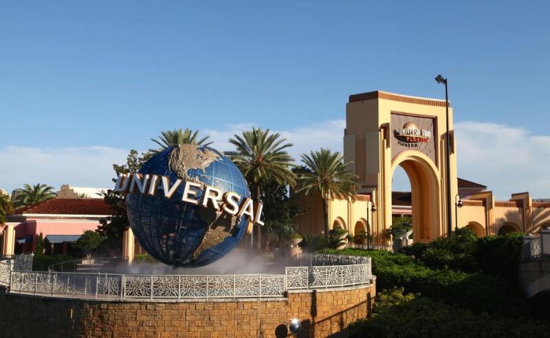 Cổng vào Universal Studios nổi bật với quả cầu xoay biểu tượng có dòng chữ 'Universal'. Bao quanh là những hàng cây cọ và thảm thực vật xanh tươi, dưới bầu trời xanh trong vắt.