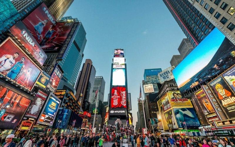 Quảng trường Times Square rực rỡ sắc màu với những biển quảng cáo sống động và dòng người tấp nập. Những tòa nhà chọc trời sừng sững phía sau dưới bầu trời trong xanh, tạo nên một bầu không khí đô thị tràn đầy năng lượng.