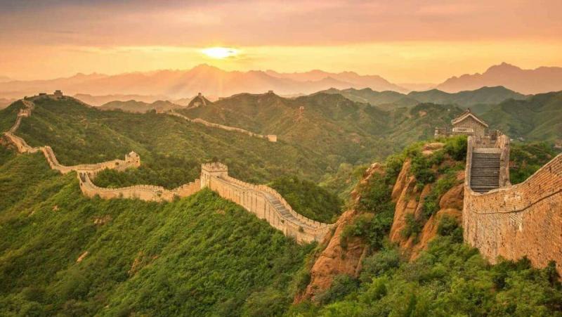Hình ảnh từ trên cao cho thấy Vạn Lý Trường Thành uốn lượn trên những ngọn đồi xanh mướt lúc hoàng hôn, với bầu trời hùng vĩ và ánh mặt trời dịu nhẹ phía sau.