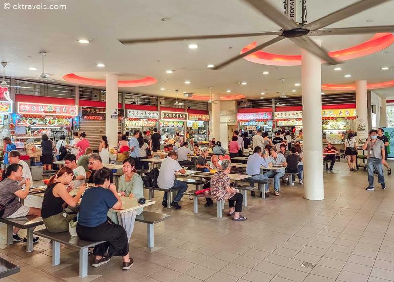 Một khu ẩm thực nhộn nhịp với nhiều quầy hàng bán đồ ăn, biển hiệu rực rỡ và nhiều người ngồi ở bàn thưởng thức bữa ăn. Không khí sống động và thân thiện.