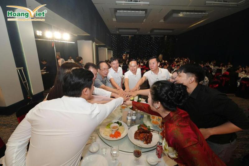 Một nhóm người đứng quanh bàn tiệc trong một căn phòng thiếu ánh sáng, mỉm cười và nắm tay nhau thể hiện sự đoàn kết. Những đĩa thức ăn được bày biện đẹp mắt, tạo nên một bầu không khí lễ hội.