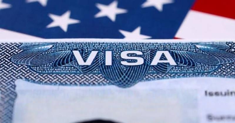 Tờ visa Mỹ cận cảnh với chữ VISA in nổi bật trên nền hoa văn màu xanh, phía sau là quốc kỳ Mỹ