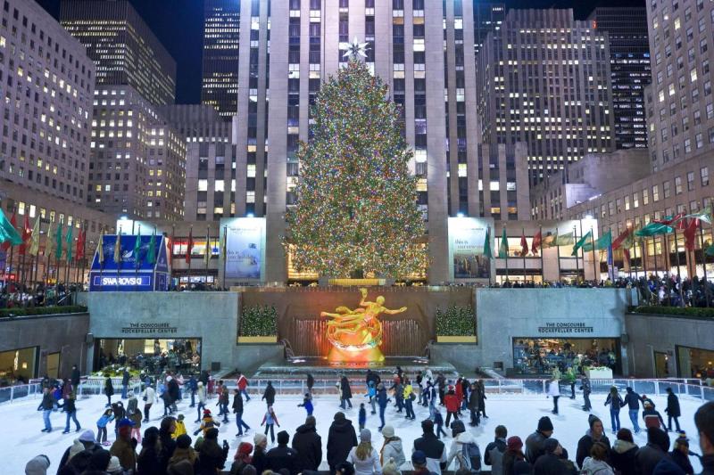 Khung cảnh lễ hội tại Rockefeller Center với một cây thông Noel lớn được trang trí lộng lẫy phía trên sân trượt băng, và mọi người đang vui vẻ trượt băng bên dưới vào ban đêm.