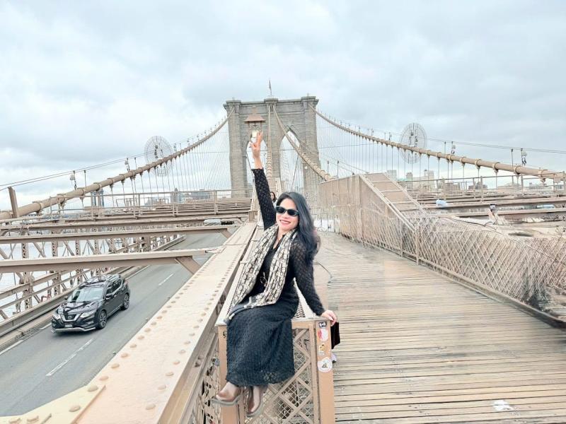 Một người phụ nữ mặc váy đen và đeo kính râm ngồi trên lan can cầu Brooklyn, mỉm cười và giơ tay lên. Phía sau là những tòa tháp và dây cáp mang tính biểu tượng của cây cầu.