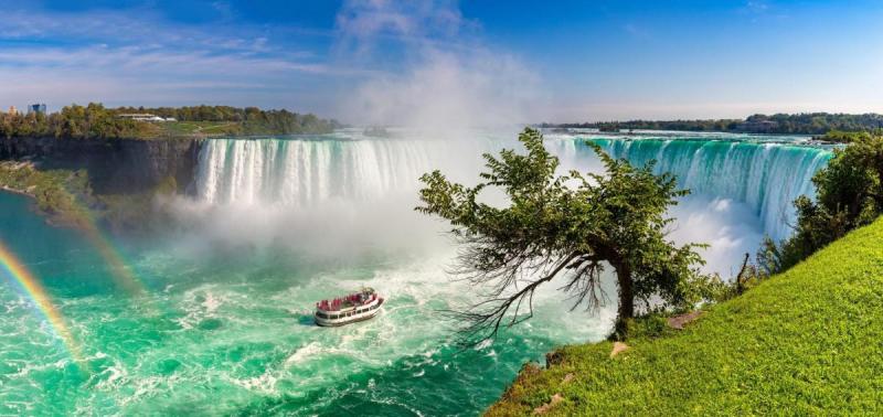 Một khung cảnh sống động của thác Niagara với một chiếc thuyền du lịch đang lướt trên mặt nước màu ngọc lam mờ ảo. Cầu vồng uốn cong ở bên trái, và thảm thực vật xanh tươi trải dài dọc theo bờ bên phải.