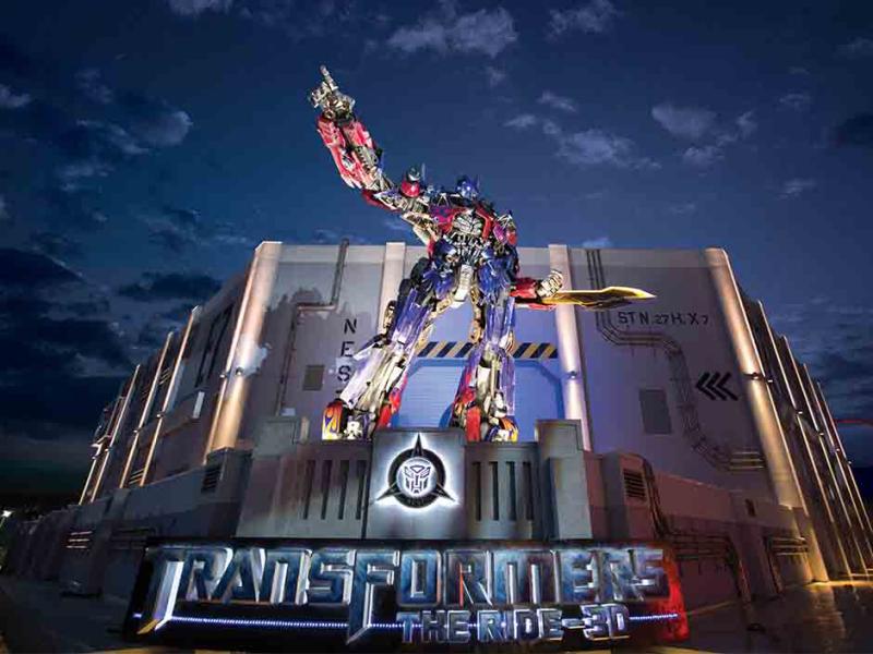 Optimus Prime, một người máy khổng lồ, đứng oai vệ trước một tòa nhà lúc hoàng hôn, chỉ tay lên trời. Biển hiệu ghi "Transformers The Ride 3D".