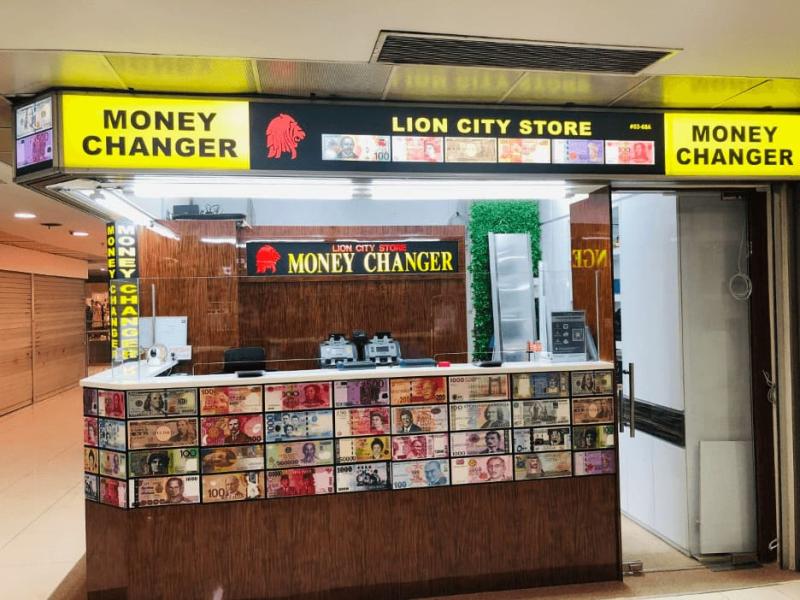 Một quầy đổi tiền mang tên "Lion City Store" trưng bày nhiều loại tiền giấy quốc tế. Biển hiệu và ánh đèn sáng tạo nên không gian chuyên nghiệp.