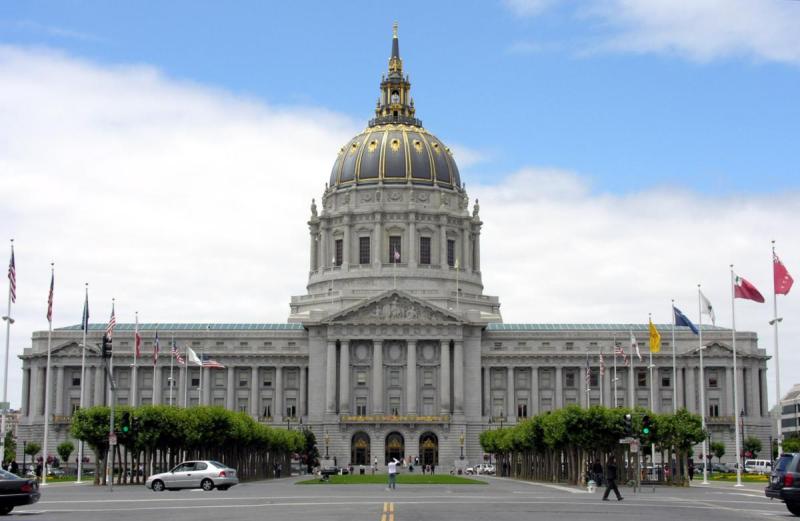 Tòa nhà thị chính ở San Francisco phong cách tân cổ điển tráng lệ với mái vòm lớn nằm dưới bầu trời nhiều mây. Cờ được treo dọc lối vào, và nhiều người đi lại gần đó.