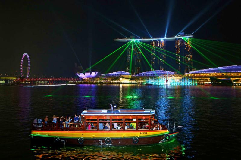 Tour singapore 4 ngày 3 đêm: Một khung cảnh buổi tối sống động tại Vịnh Marina của Singapore, với ánh đèn laser đầy màu sắc từ Marina Bay Sands, một chiếc thuyền ở phía trước và một vòng đu quay.