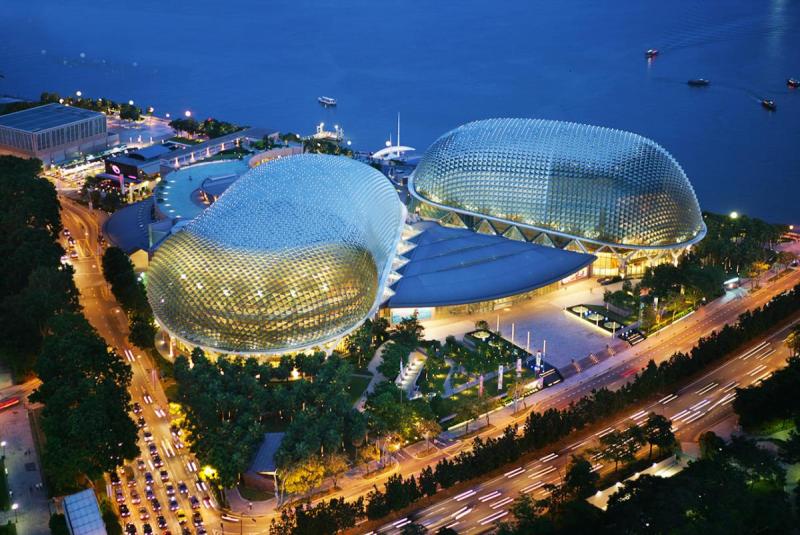 Hình ảnh từ trên cao cho thấy các rạp chiếu phim Esplanade ở Singapore vào ban đêm, với những cấu trúc mái vòm phát sáng bên bờ nước, được bao quanh bởi ánh đèn thành phố và đường phố.