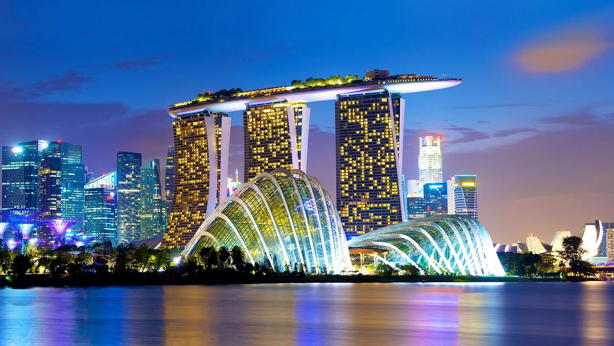 Khung cảnh đường chân trời của Singapore lúc hoàng hôn với Marina Bay Sands và những công trình kiến trúc hiện đại được chiếu sáng. Bầu trời xanh ngắt phản chiếu trên mặt nước tĩnh lặng, tạo nên một khung cảnh sống động và hiện đại.