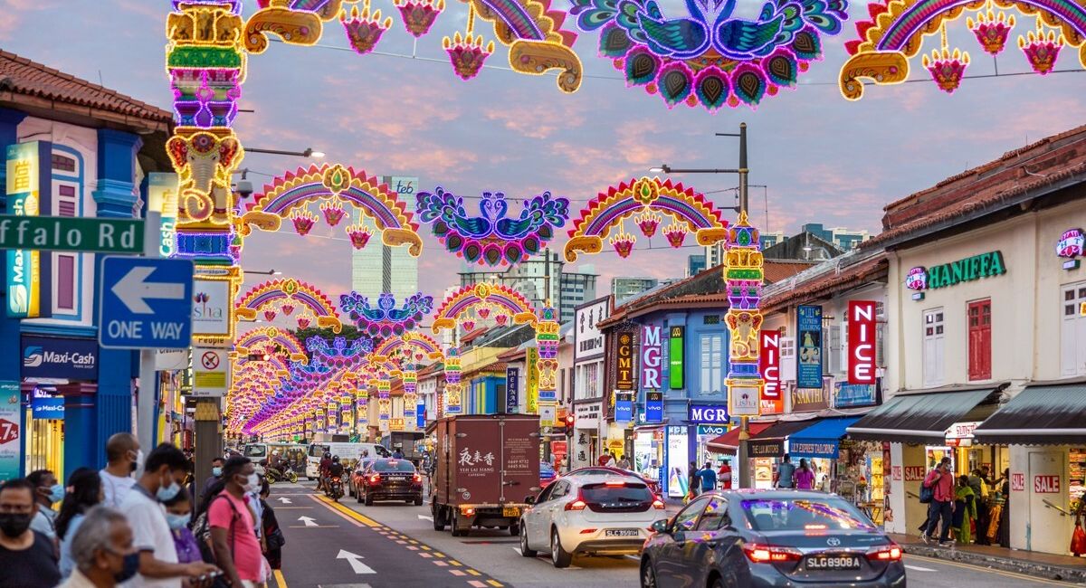 Cảnh đường phố ở khu Little India, Singapore, lúc hoàng hôn với những ánh đèn lễ hội rực rỡ uốn lượn phía trên. Các tòa nhà nằm dọc theo con phố, và người đi bộ cùng xe cộ tấp nập khắp nơi.