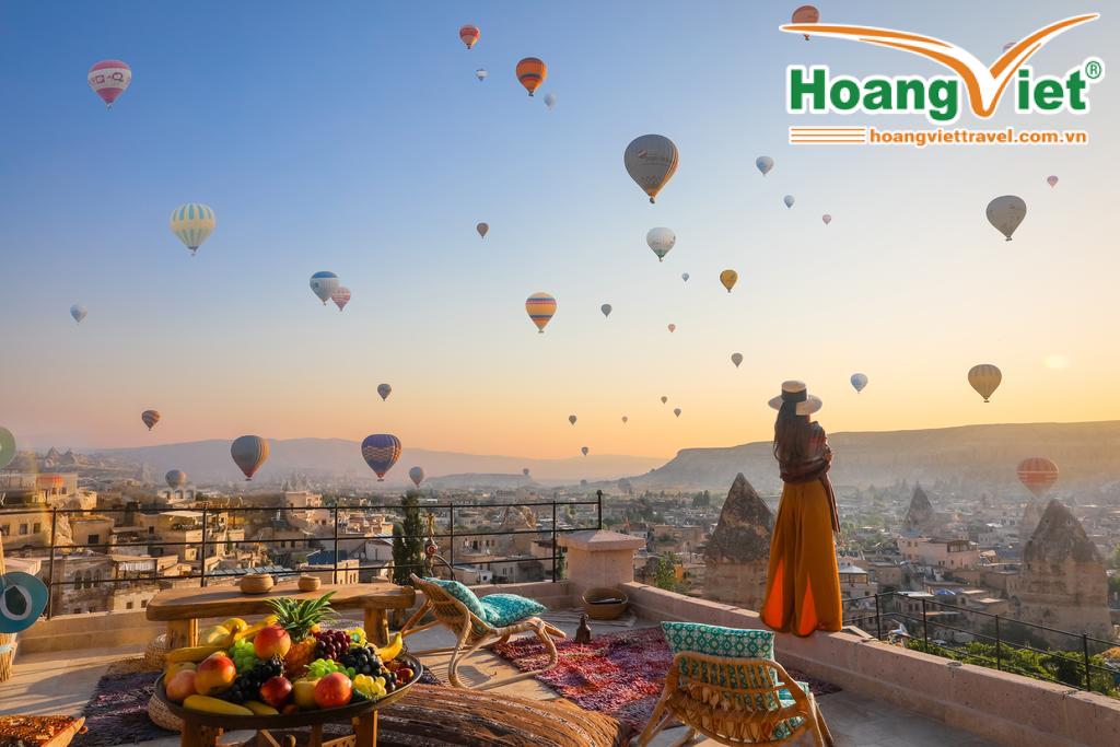 Một người phụ nữ trong bộ váy dài thướt tha và đội mũ đang chiêm ngưỡng vô số khinh khí cầu bay lơ lửng trên bầu trời Cappadocia lúc bình minh. Phía trước là một sân hiên đầy màu sắc với trái cây và ghế ngồi.