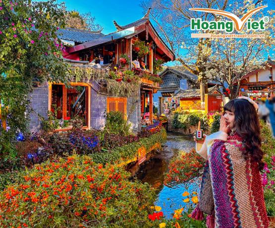 Một người phụ nữ quấn khăn choàng nhiều màu sắc chụp ảnh tự sướng trước một khu vườn rực rỡ và một tòa nhà truyền thống châu Á bên bờ suối, dưới bầu trời xanh trong vắt.