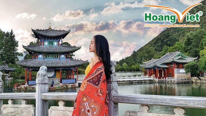 Cô gái khoác khăn họa tiết đỏ đứng bên cầu đá trước lầu Đắc Nguyệt ở hồ Hắc Long Đầm Lệ Giang