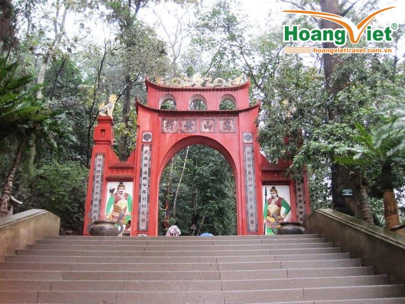 Tour Du Lịch Đền Hùng Cổng vòm màu đỏ được trang trí công phu với các ký tự châu Á ở phía trên, hai bên là những hàng cây cao. Cầu thang dẫn lên trên, với những bức tranh tường đầy màu sắc và phông nền là khu rừng tươi tốt.