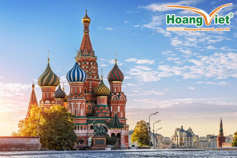 Cảnh tượng Nhà thờ Thánh Basil rực rỡ sắc màu ở Moscow dưới ánh nắng mặt trời, với những mái vòm hình củ hành đầy màu sắc, được bao quanh bởi cây cối và bầu trời trong xanh với những đàn chim bay lượn, tạo nên một bầu không khí thanh bình.