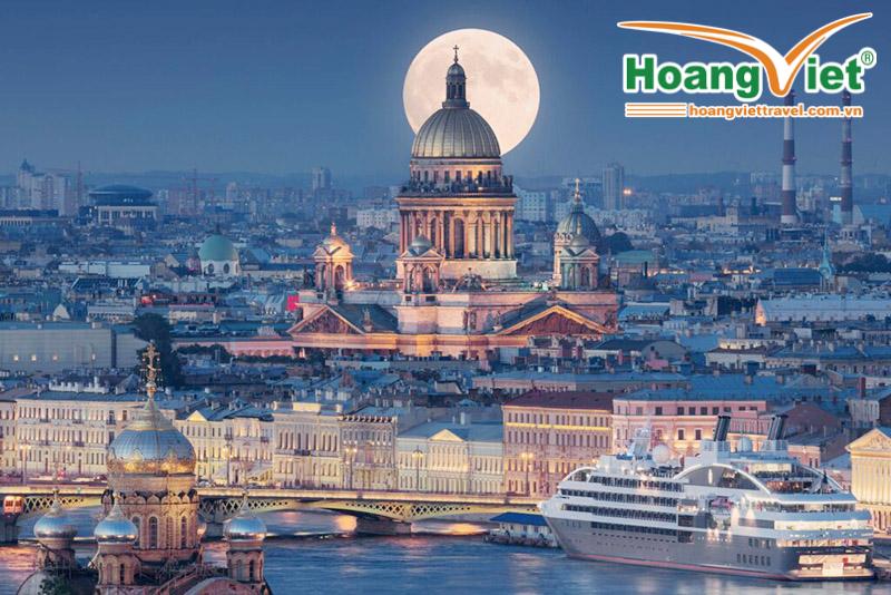 Trăng tròn tỏa sáng phía sau một nhà thờ lớn được thắp sáng rực rỡ giữa khung cảnh thành phố rộng lớn. Một du thuyền neo đậu gần những tòa nhà trang trí công phu bên bờ sông, toát lên vẻ sang trọng.