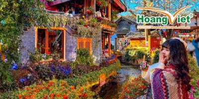 tour lệ giang - shangrila Một người phụ nữ quấn khăn choàng nhiều màu sắc chụp ảnh tự sướng trước một khu vườn rực rỡ và một tòa nhà truyền thống châu Á bên bờ suối, dưới bầu trời xanh trong vắt.