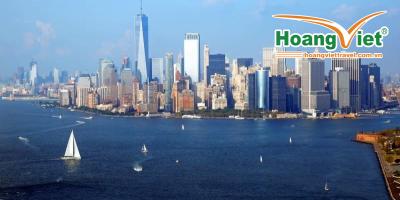 Toàn cảnh thành phố New York với những tòa nhà chọc trời hiện đại dưới bầu trời xanh trong vắt. Những chiếc thuyền buồm lướt nhẹ trên mặt nước tĩnh lặng, tạo nên một khung cảnh thành phố thanh bình và sống động.