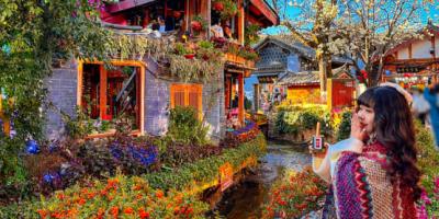 tour lệ giang - shangrila Một người phụ nữ quấn khăn choàng nhiều màu sắc chụp ảnh tự sướng trước một khu vườn rực rỡ và một tòa nhà truyền thống châu Á bên bờ suối, dưới bầu trời xanh trong vắt.
