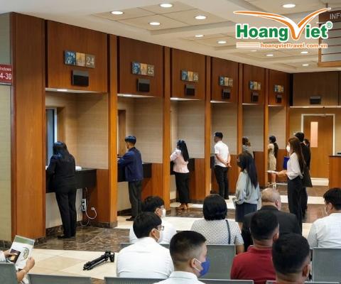 Mọi người xếp hàng tại các quầy dịch vụ trong một văn phòng sáng sủa, hiện đại. Khách hàng, một số ngồi và một số chờ đợi, tạo nên một bầu không khí yên tĩnh, ngăn nắp.
