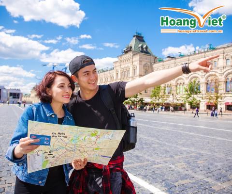 Một cặp đôi tươi cười, tay cầm bản đồ, đứng trong một quảng trường rực rỡ ánh nắng với những tòa nhà cổ kính. Người đàn ông chỉ tay về phía trước, như muốn chỉ hướng hoặc gợi lên điều gì đó thú vị.
