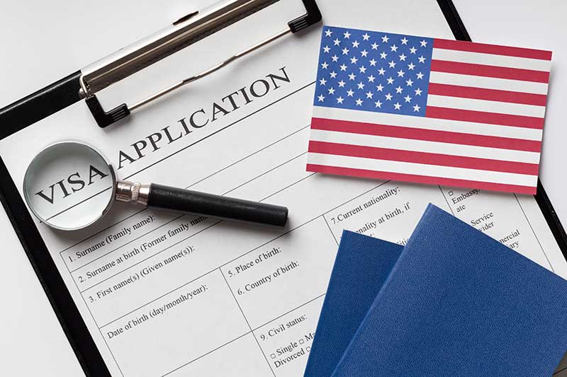 Mẫu đơn xin visa được kẹp trên bảng kẹp giấy cùng với một kính lúp, một lá cờ Mỹ và hai cuốn hộ chiếu màu xanh lam, tượng trưng cho việc đi lại hoặc nhập cư.