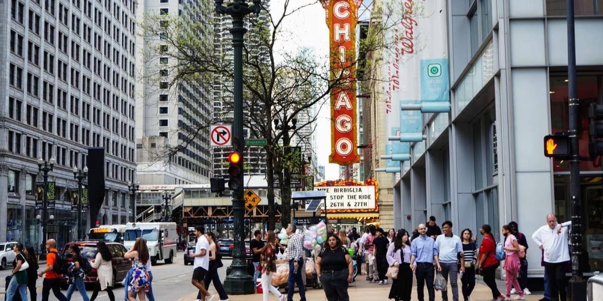 Cảnh đường phố nhộn nhịp ở Chicago với đủ loại người đi bộ và xe cộ trước biển hiệu nổi tiếng của Nhà hát Chicago. Không khí sống động và đậm chất đô thị.