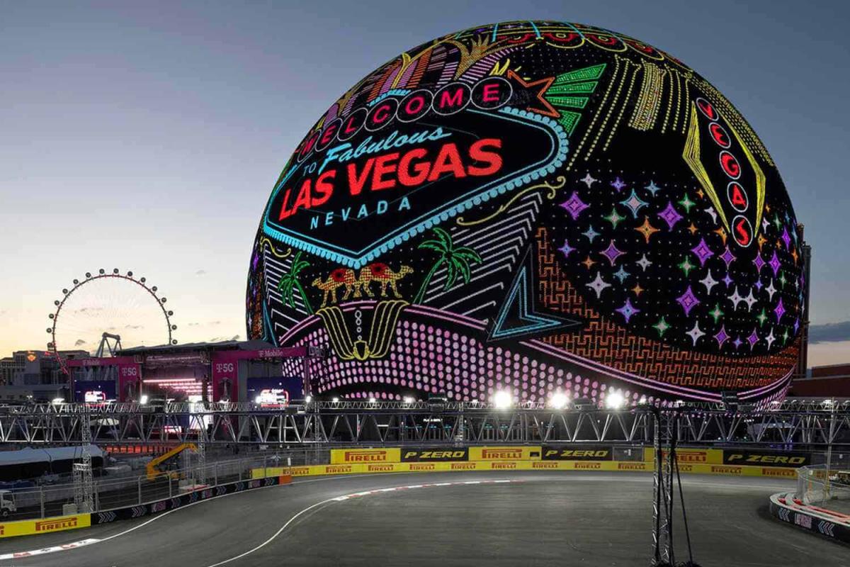 Quả cầu phát sáng Las Vegas hiển thị dòng chữ "Chào mừng đến với Las Vegas tuyệt vời" bằng màu neon rực rỡ. Vòng đu quay và đường đua hiện ra lúc hoàng hôn. Không khí tràn đầy năng lượng.