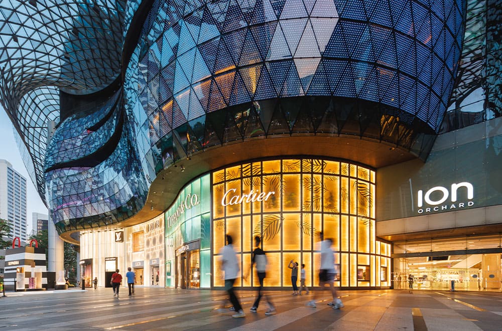 Mặt tiền kính hiện đại của trung tâm mua sắm ION Orchard lúc hoàng hôn. Ánh sáng ấm áp từ các cửa hàng sang trọng, bao gồm cả Cartier, và hình ảnh những người mua sắm đi ngang qua mờ ảo.