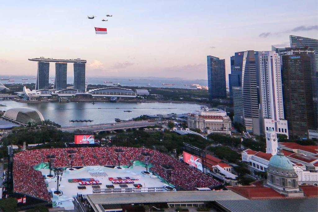 Ảnh chụp từ trên cao lễ duyệt binh Quốc khánh Singapore tại vịnh Marina. Máy bay trực thăng mang theo một lá cờ lớn. Có thể nhìn thấy các tòa nhà chọc trời và một nhà hát ngoài trời với đám đông khán giả.