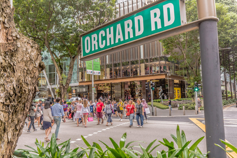 Khung cảnh nhộn nhịp trên đường Orchard Road cho thấy mọi người băng qua đường tại vạch kẻ dành cho người đi bộ, xung quanh là cây xanh tươi tốt và các cửa hàng cao cấp như Gucci. Không khí nơi đây thật sống động và náo nhiệt.