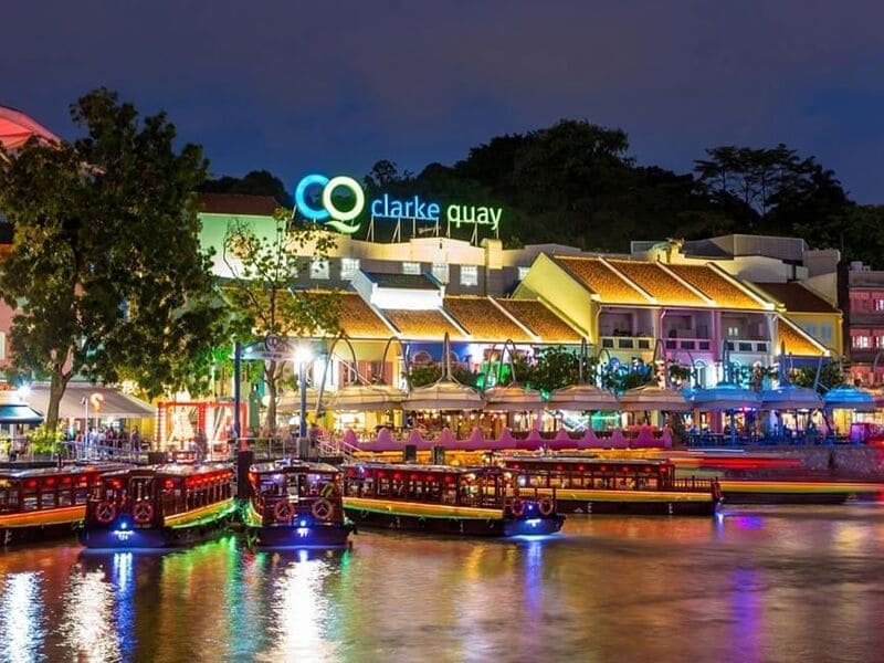 Cảnh đêm lung linh tại Clarke Quay, Singapore. Ánh đèn rực rỡ chiếu sáng các quán bar và nhà hàng ven sông, cùng những chiếc thuyền neo đậu trên dòng sông yên bình. Không khí tràn đầy năng lượng.