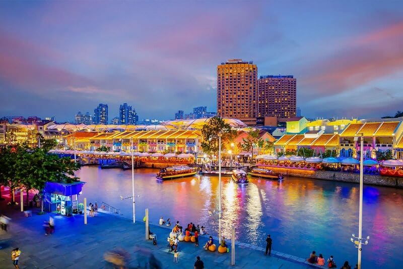 Khung cảnh ven sông rực rỡ sắc màu lúc hoàng hôn, với những tòa nhà và thuyền bè được chiếu sáng trên mặt nước, mọi người thư giãn trên lối đi dạo, cùng với phông nền là một thành phố sống động.
