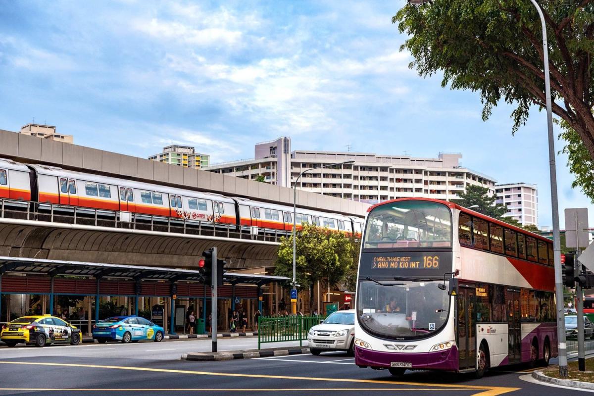 Một con phố đô thị nhộn nhịp với xe buýt hai tầng đang chạy qua, taxi chờ sẵn và đường ray xe lửa trên cao ở phía xa. Bầu trời xanh và cây cối càng làm tăng thêm vẻ sống động.