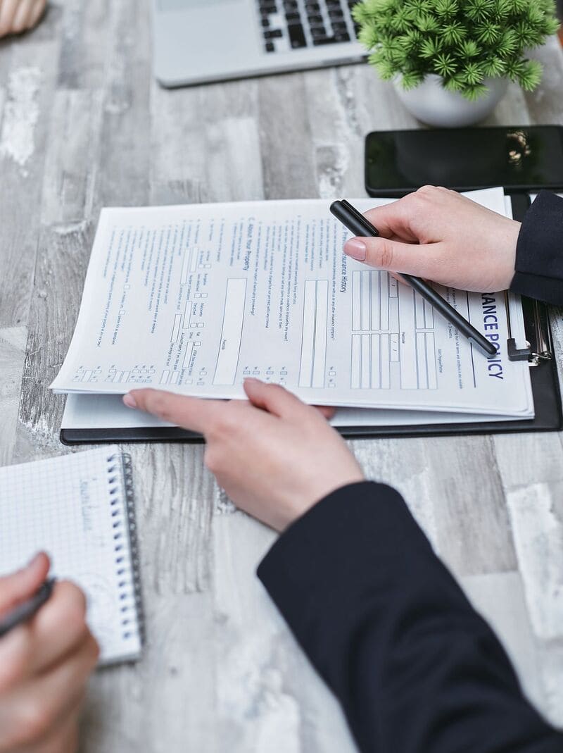 Bàn tay cầm bút chỉ vào tờ hợp đồng bảo hiểm Insurance Policy kẹp trên bảng trình ký đặt trên bàn làm việc