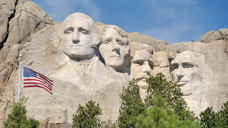Dưới bầu trời xanh, tượng đài Mount Rushmore khắc bốn khuôn mặt tổng thống được tạc trên đá granit. Lá cờ Mỹ tung bay bên trái, bao quanh là những hàng cây xanh.